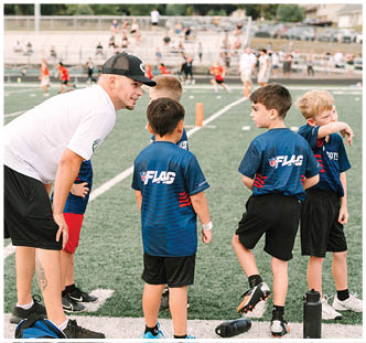 An adult wearing a white shirt and black cap is crouched down talking to a group of children in blue sports jerseys standing on a football field. Description generated by AI