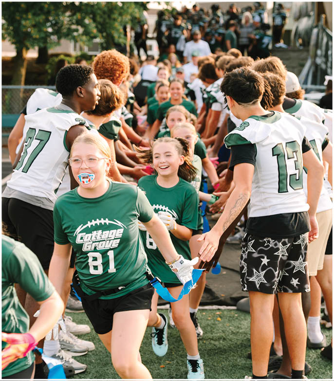 Children wearing sports uniforms and casual athletic clothing are running through a tunnel formed by people giving high fives on a sports field. Description generated by AI