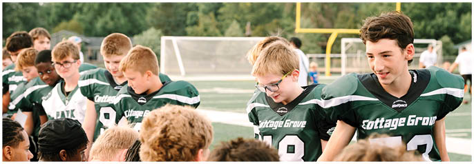 A group of children wearing green and white Cottage Grove football uniforms are gathered on a sports field, some smiling and interacting with each other. Description generated by AI