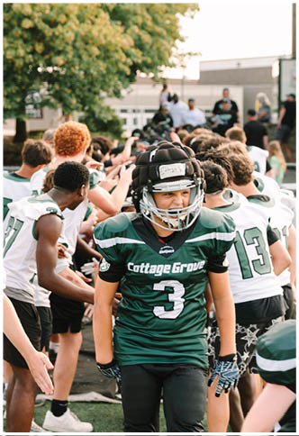 A person in a green Cottage Grove football uniform and helmet walks past a line of people giving high-fives on a sports field. Description generated by AI