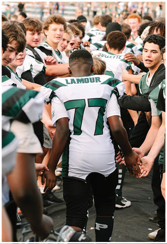 A group of people in sports uniforms are gathered closely together, with one person wearing a white jersey labeled “LAMOUR 77" walking through and giving high-fives. Description generated by AI