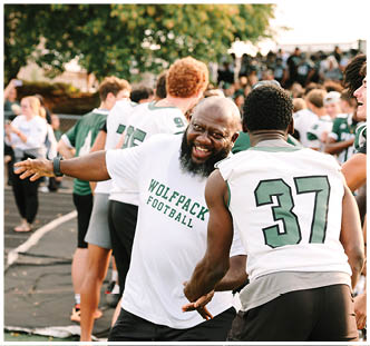 A person wearing a white Wolfpack Football shirt is smiling and reaching out to another person in a sports jersey with the number 37 on the back, with several other people and a blurred background of trees and spectators. Description generated by AI