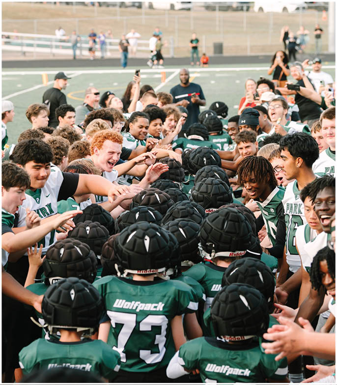 A group of people in sports jerseys enthusiastically reach out to high-five a team of young athletes wearing green Wolfpack uniforms and padded helmets on a football field. Description generated by AI