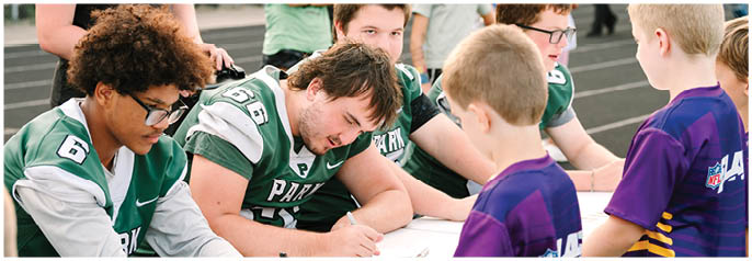 Several people wearing green sports jerseys are seated at a table signing autographs for children dressed in purple shirts outdoors. Description generated by AI