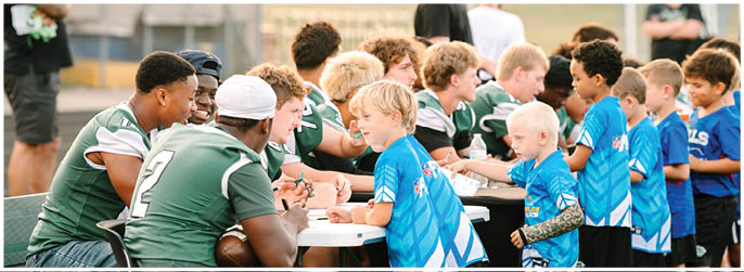 Children in blue sports uniforms are shaking hands and interacting with adults in green sports uniforms across a table outdoors. Description generated by AI