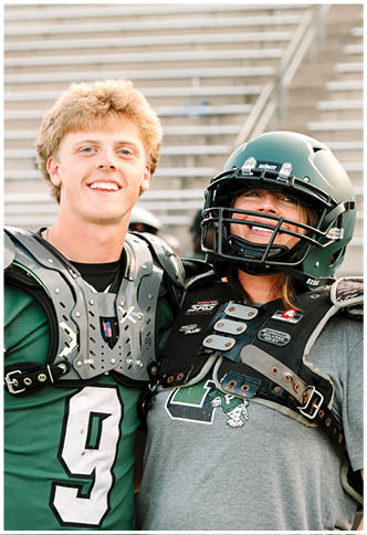 Two people wearing football shoulder pads stand closely together on bleachers, one smiling with short curly hair and the other wearing a helmet and a gray shirt. Description generated by AI