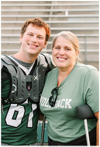 A person wearing a football uniform with shoulder pads and a jersey numbered 61 is standing next to another person in a green shirt with sunglasses hanging from the collar, both smiling in an outdoor stadium setting. Description generated by AI