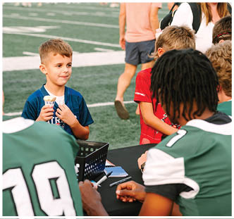 A child holding an ice cream cone stands in front of two people in sports jerseys seated at a table on a sports field. Description generated by AI