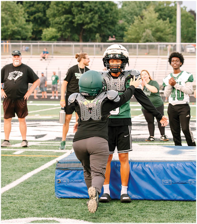Two people wearing football helmets and shoulder pads are interacting on a sports field while several others watch in the background. Description generated by AI