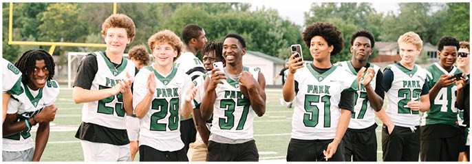A group of people wearing matching sports jerseys stand together on a field, smiling, clapping, and holding a phone. Description generated by AI