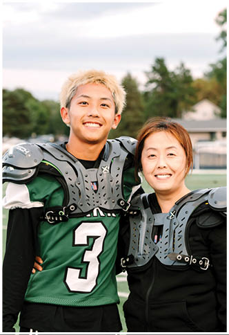 A person wearing a green football jersey with the number three and shoulder pads is smiling on a sports field next to another person partially visible in similar gear. Description generated by AI
