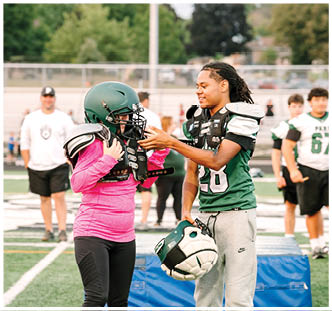 Two people on a sports field are putting on football helmets and shoulder pads while another person stands in the background. Description generated by AI