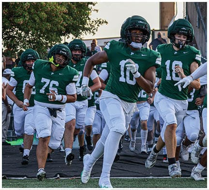 People in green football uniforms and helmets are running onto a field from a tunnel area near bleachers with a tree and mascot visible in the background. Description generated by AI