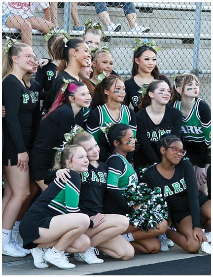 A group of adults in matching black and green cheerleading uniforms with bows in their hair pose together outdoors near bleachers. Description generated by AI