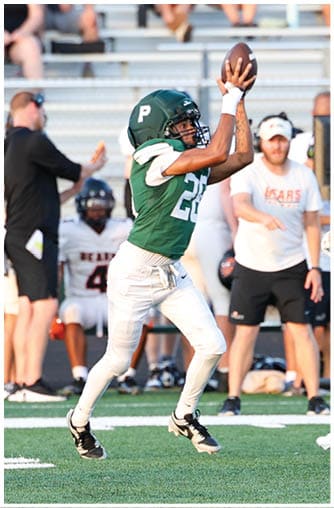 A person in a green football uniform is jumping to catch a football on a field while other players and people stand in the background. Description generated by AI