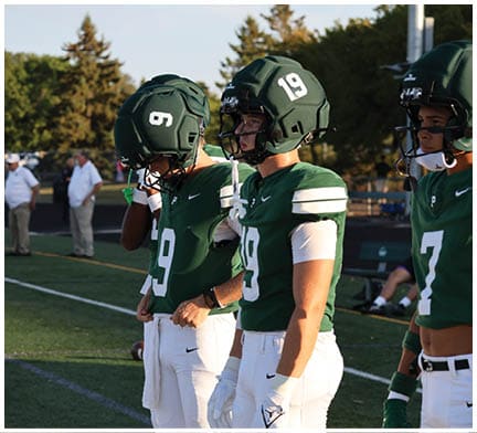 Three people wearing green football uniforms and helmets stand on a field with trees and other people in the background. Description generated by AI