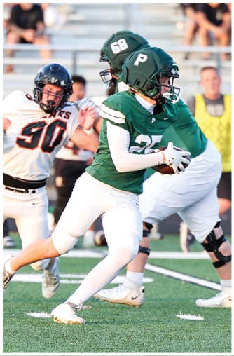 People wearing green and white football uniforms are playing on a field with one person holding the ball and others preparing to block or run. Description generated by AI