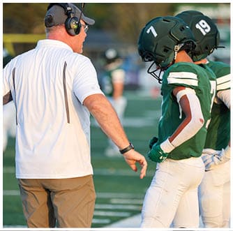 An adult wearing a white shirt and brown pants is reaching out toward a person in a green sports jersey and white pants standing on a field. Description generated by AI