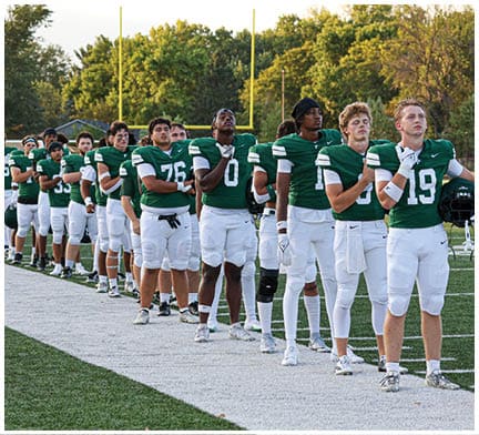 A line of people in green and white football uniforms stand on a field with their hands over their hearts, facing forward near goalposts and trees in the background. Description generated by AI