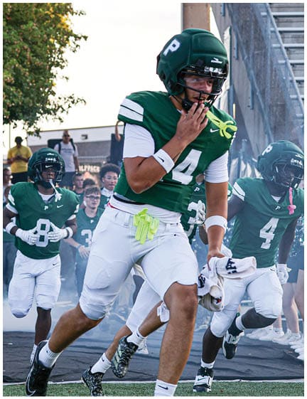 Football players in green and white uniforms are running onto the field from a tunnel with cheerleaders and a mascot on either side. Description generated by AI