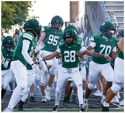 A group of people wearing green and white football uniforms and helmets are running together on a field near bleachers and trees. Description generated by AI