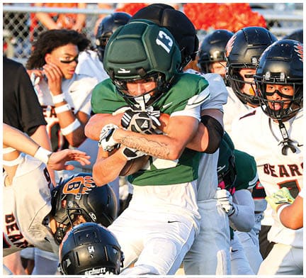 A group of people wearing football uniforms are actively engaged in a play on a football field with one person holding the ball tightly while others attempt to tackle or block. Description generated by AI
