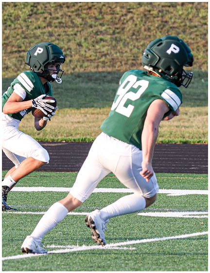 Two people wearing green football helmets and jerseys are running on a football field, with one holding a football. Description generated by AI