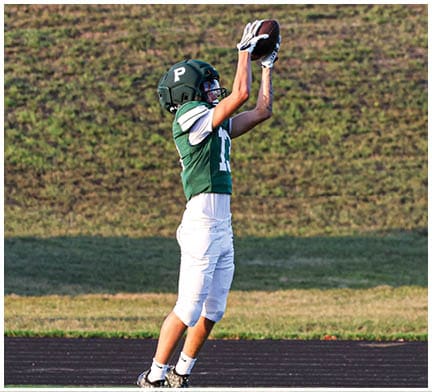 Two people in green and white football uniforms are on a field, with one jumping to catch a football while the other runs nearby. Description generated by AI