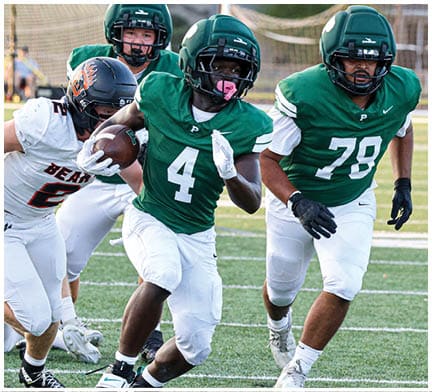 A group of people in football uniforms are actively playing on a field with one person in a green jersey running with the ball while others in white jerseys attempt to tackle or block. Description generated by AI
