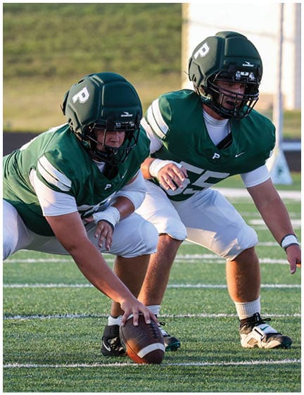Two people in green football uniforms and helmets are positioned on a football field, with one person holding a football ready to snap it. Description generated by AI