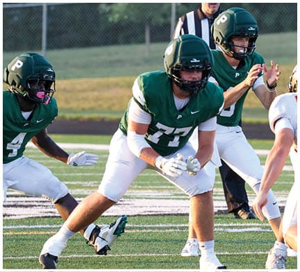 Four people in green and white football uniforms and one person in a white and black football uniform are on a field with a referee in the background. Description generated by AI