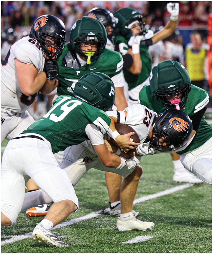 Several football players in green and white uniforms tackle a player in a white uniform holding the ball during a game on a grassy field. Description generated by AI