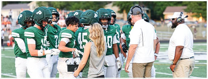 A group of football players in green uniforms and helmets are gathered on a field with two adults in white shirts and headsets standing nearby. Description generated by AI