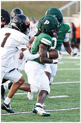 A person wearing a green football uniform with a helmet is running with a football while being pursued by opponents in white uniforms on a grassy field. Description generated by AI