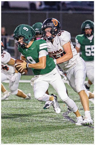 Two people in football uniforms are engaged in a play on a field, with one person in green running while holding the ball and another in white attempting to tackle them. Description generated by AI
