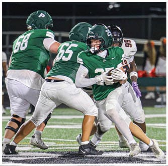 People wearing football uniforms are engaged in a play on a field with artificial turf and yard lines visible. Description generated by AI