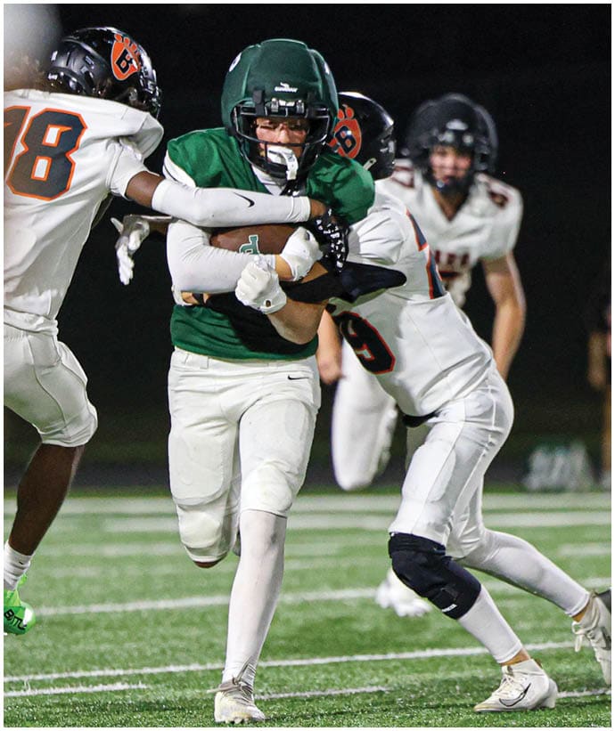 A person in a green football uniform is running with the ball while two people in white uniforms attempt to tackle them on a football field at night. Description generated by AI