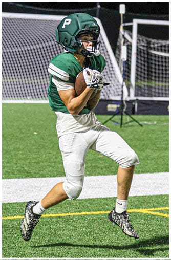 Two people in football uniforms are on a field with artificial turf and white and yellow lines, with one person reaching out toward the other. Description generated by AI