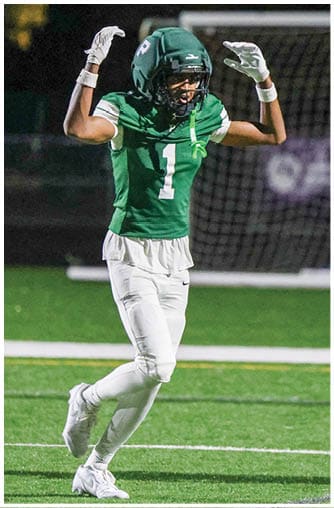 A person wearing a green football jersey with the number one and white pants is raising their arms on a football field at night. Description generated by AI