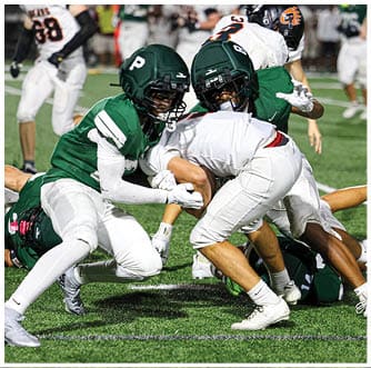 Several people in white sports uniforms are engaged in a close physical play on a green turf field, with one person in a dark green uniform on the ground. Description generated by AI