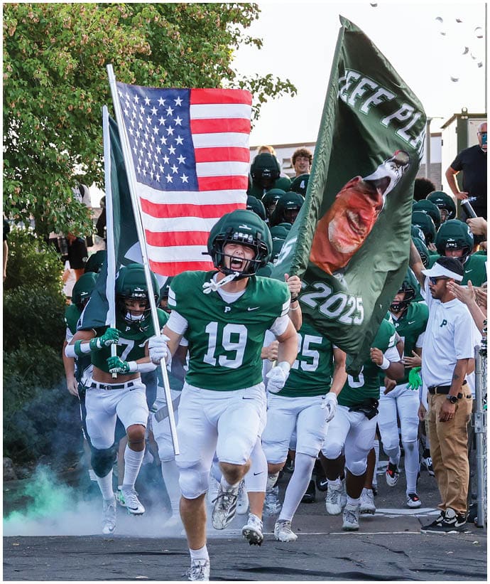 A group of people in green and white football uniforms run energetically onto a field while holding an American flag and a large green flag with a face and text, surrounded by cheering spectators. Description generated by AI