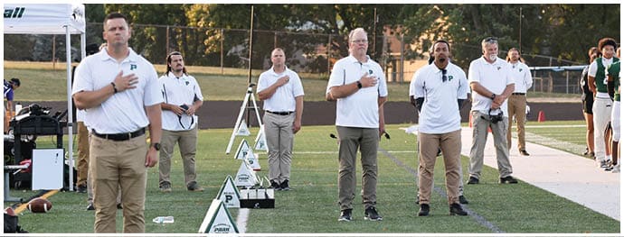Adults wearing white polo shirts and khaki pants stand on a football field with their hands over their hearts, facing forward in a line near a sideline. Description generated by AI