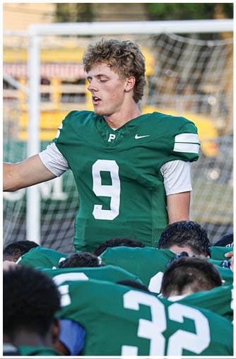 A person wearing a green football jersey with the number nine stands among a group of people in green jerseys who are bent over, with a goal net visible in the background. Description generated by AI