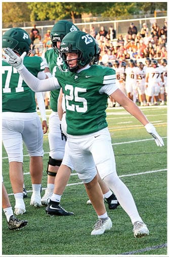People wearing green and white football uniforms are standing on a field, with one person raising an arm and wearing a helmet and gloves. Description generated by AI