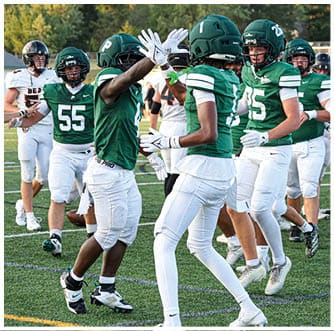 People wearing green jerseys and white football pants are standing and moving on a grassy field. Description generated by AI