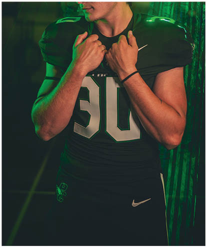 A person wearing a football uniform with the number 30 and a cowboy hat stands with hands near the collar, looking down against a dark background with green lighting. Description generated by AI