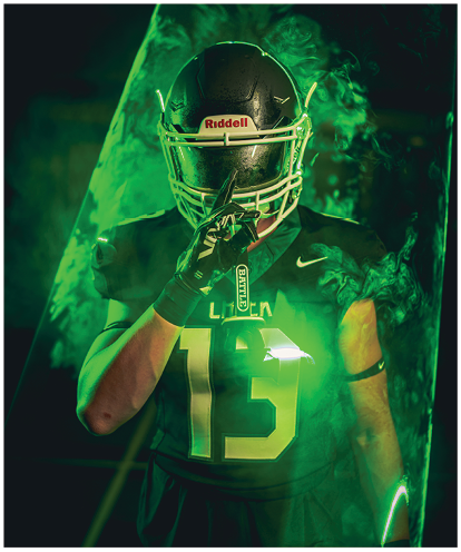 A person wearing a dark football uniform with the number 65 holds a brightly colored striped flag behind them with dramatic lighting and a dark background. Description generated by AI
