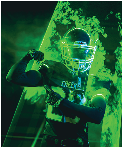 A person wearing a football uniform with the word Creek and the number 23 flexes one arm and points with the other while surrounded by green smoke and glowing green light effects. Description generated by AI