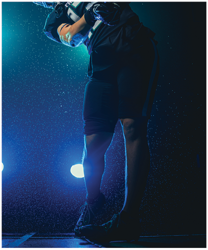 A person wearing sports gear and a helmet is swinging a golf club under bright stadium lights against a dark background. Description generated by AI