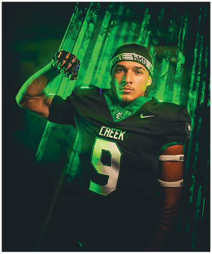 A person wearing a black football uniform with the number nine and the word Creek flexes their arm while standing against a green-lit background. Description generated by AI